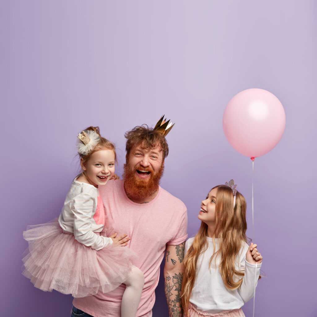 Vertical shot of overemotive joyful single dad and two daughters, celebrate Fathers Day, wear festive outfits, have fun together, isolated over purple background, blank space above for promotion