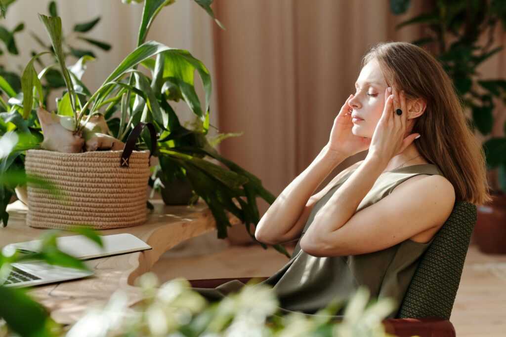 A woman relaxes, massaging her temples among indoor plants in a bright, sunlit room.