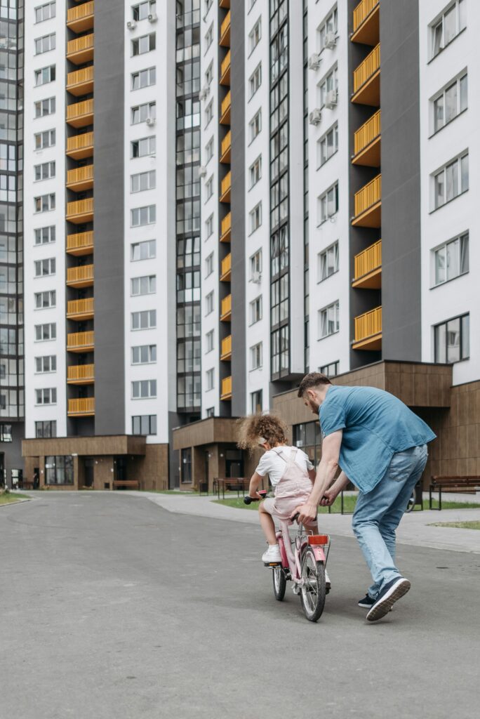 A father guides his daughter on a bike ride near towering apartment buildings.